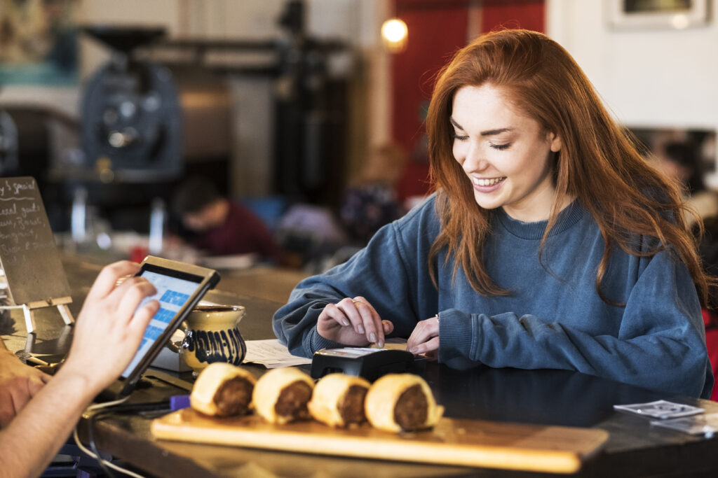 Smiling young woman with long red hair standing at a counter in a restaurant, paying her bill.